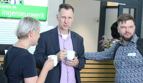 Zwei Personen im Gespräch an einem Stehtisch vor großen Fenstern.